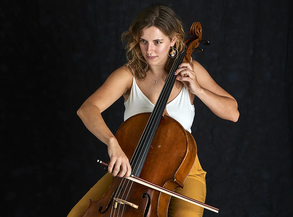 Young woman with curly blonde hair in a white sleeveless top and yellow pants, playing a cello with focus. She holds the cello with one hand and a bow with the other, against a dark background.
