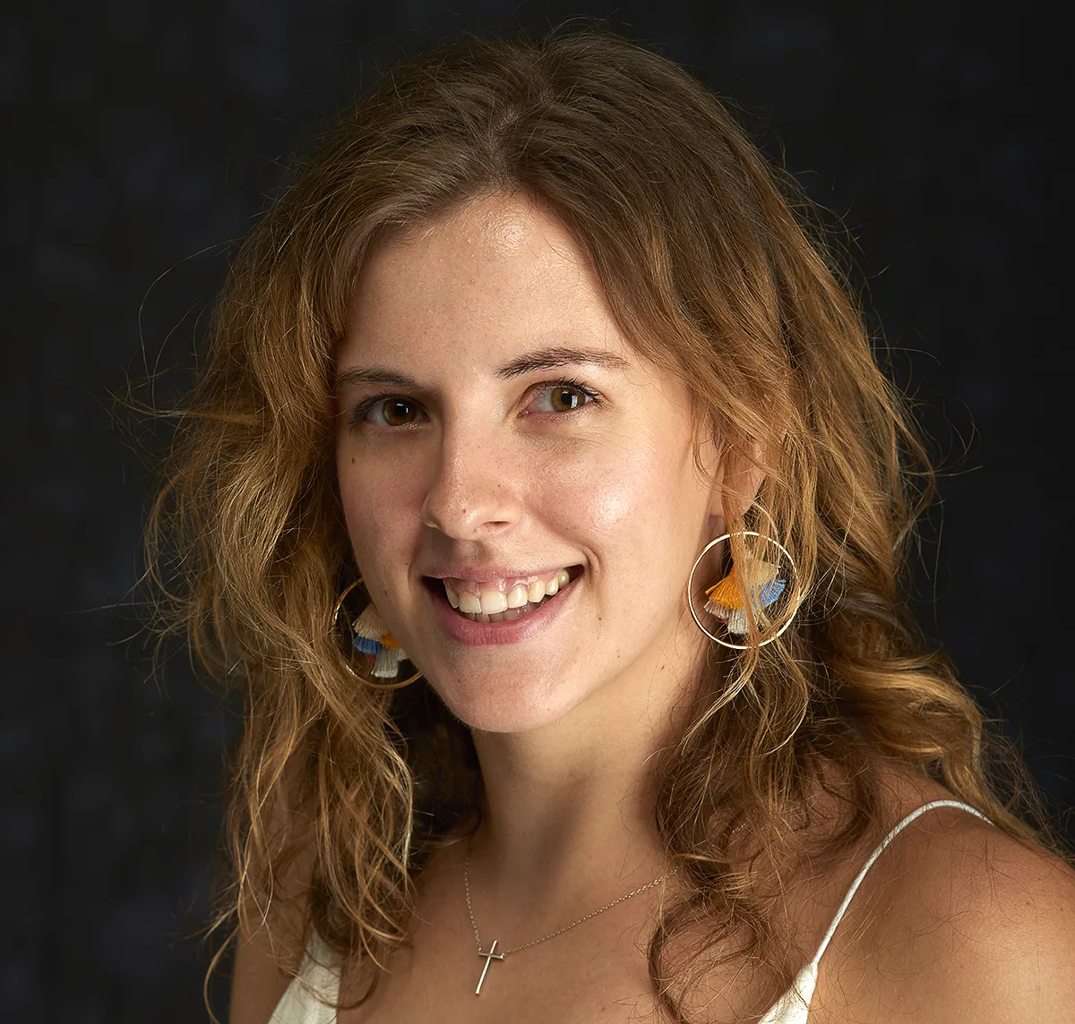 Portrait of a smiling young woman with wavy light brown hair, wearing a pair of colorful hoop earrings and a delicate cross necklace. She is dressed in a white top and is set against a dark background.