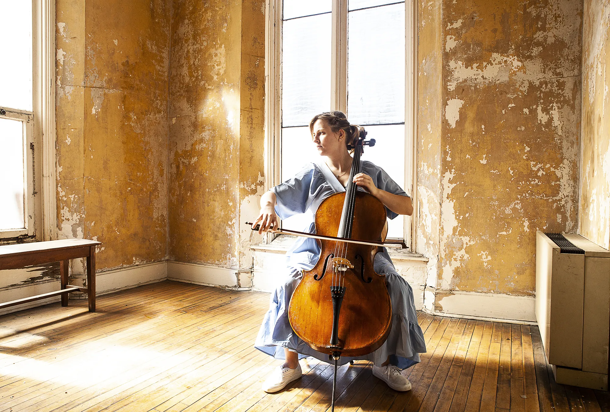 Woman with blonde hair in a light blue dress playing a cello in a sunlit room with peeling yellow walls and wooden floors. She is seated near a large window, focused on her performance, with natural light illuminating the scene. The room has a rustic, aged appearance, with a wooden bench and a heater against the walls.